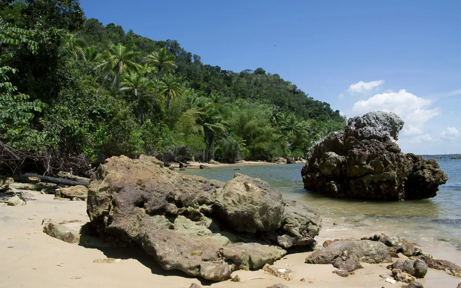 Praia da Gamboa com blocos de pedra na areia, água calma e mata fechada na encosta
