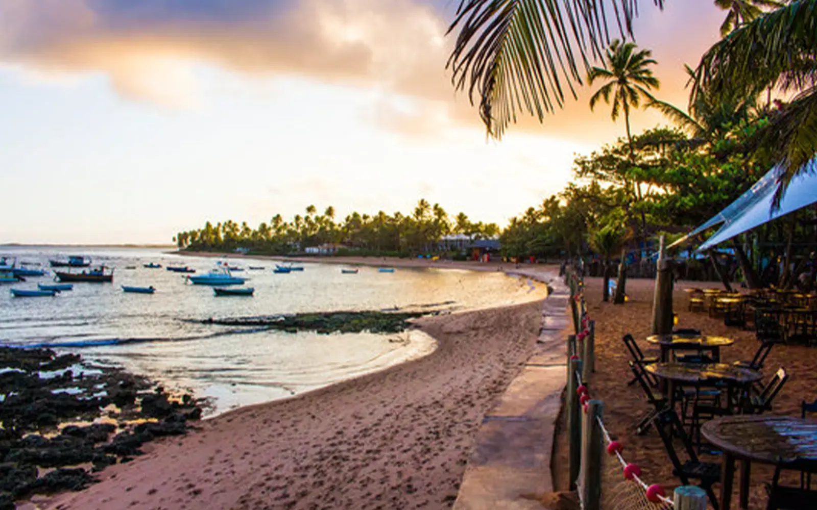 Fim de tarde na Praia do Forte, com barcos ancorados na enseada e mesas de bar sob os coqueiros