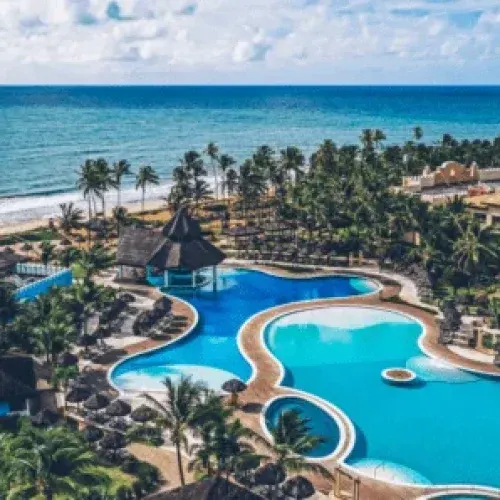 Vista aérea de um resort à beira-mar, com piscinas, coqueiros e a praia ao fundo