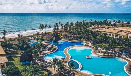Vista aérea de um resort à beira-mar, com piscinas, coqueiros e a praia ao fundo