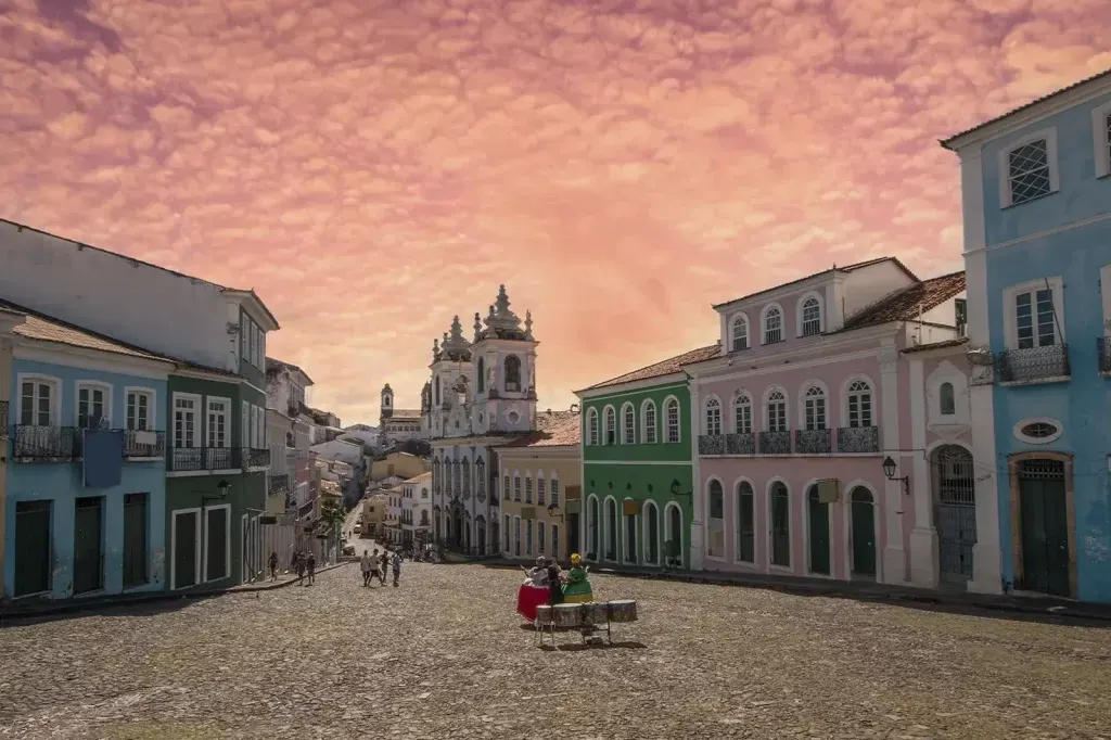 Largo do Pelourinho ao entardecer, com os casarões coloridos, a igreja ao fundo e um grupo tocando tambores
