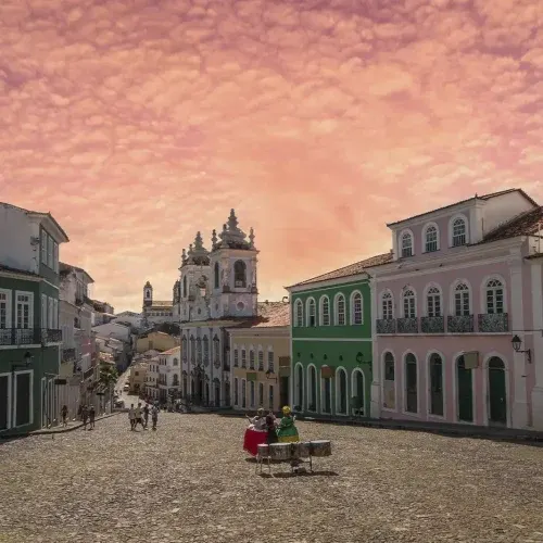 Largo do Pelourinho ao entardecer, com os casarões coloridos, a igreja ao fundo e um grupo tocando tambores
