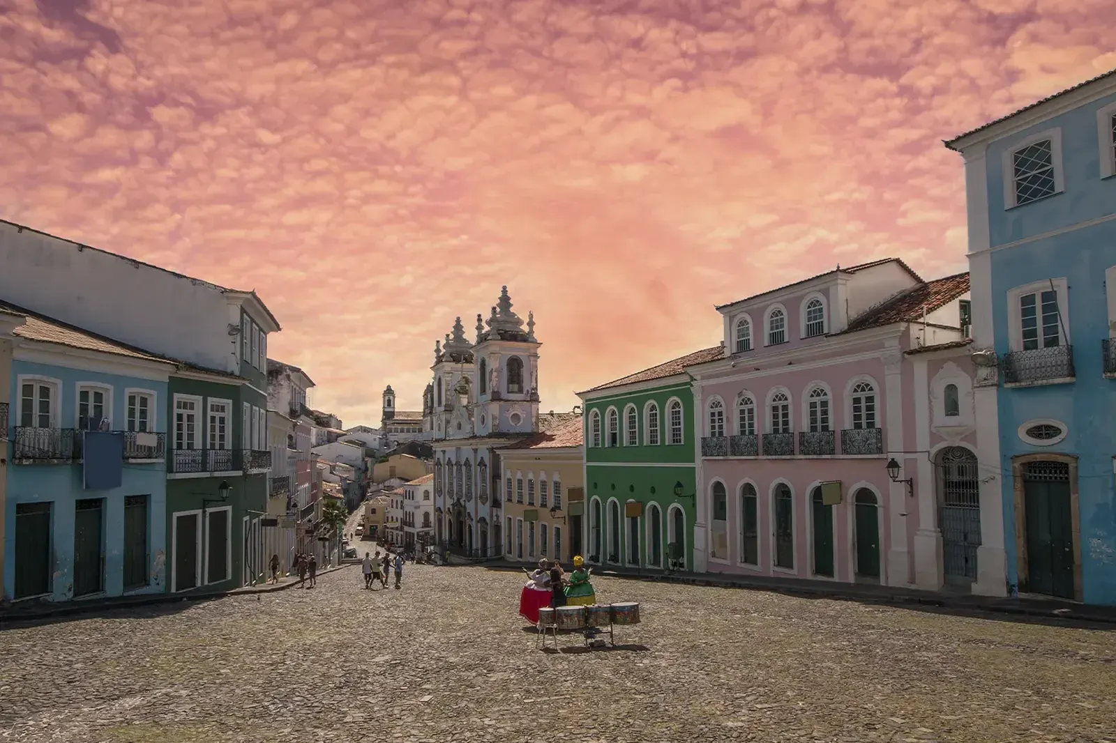 Largo do Pelourinho ao entardecer, com os casarões coloridos, a igreja ao fundo e um grupo tocando tambores